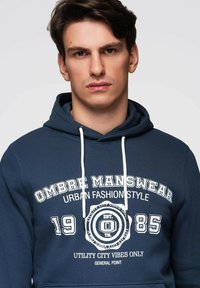 Sweat à capuche bleu marine en tissu doux, doté de motifs blancs, de cordons de serrage et d'une poche kangourou. Le design comprend du texte sur la mode urbaine et un logo rond.