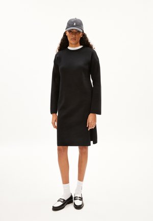 Robe noire à manches longues avec un col rond, en tissu texturé, fente sur le côté, associée à une casquette foncée et des chaussures noires avec des chaussettes blanches.