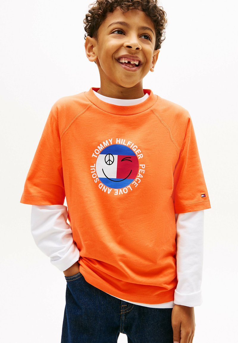 Garçon souriant aux cheveux bouclés portant un t-shirt orange Tommy Hilfiger par-dessus un manches longues blanches, les mains dans les poches, debout devant un fond blanc.