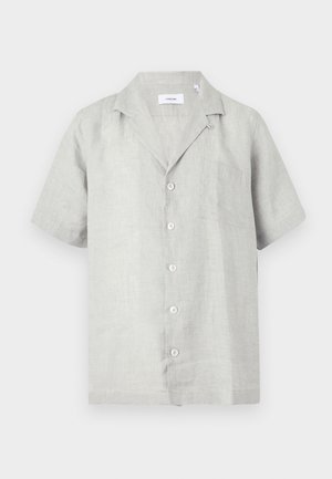 Uomo in piedi con pantaloni beige, camicia leggera a maniche corte abbottonata e scarpe eleganti marrone scuro su sfondo bianco semplice.