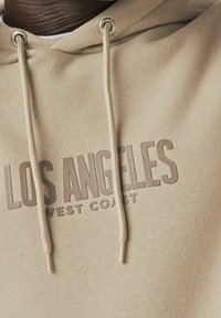 Sudadera beige con cordón, presenta letras en relieve "LOS ANGELES WEST COAST" en un tono más oscuro. Textura suave, diseño casual.