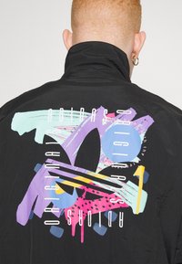 Svart vindjacka med färgstarkt motiv på ryggen, med abstrakta former i rosa, blått, lila och gult, samt vita textdetaljer.