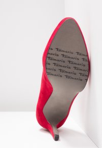 Červený semišový botík na vysokém podpatku s špičatou špičkou a štíhlým stiletto podpatkem. Na podrážce se opakovaně objevuje značka "Tamaris".