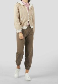 Person iført en beige lynlåsjakke, skjorte med rosa mønster, brune opsmøgede bukser og hvide sneakers, der står op ad en ensfarvet lys baggrund.