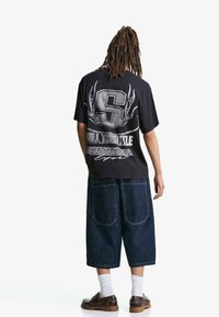 Črna oversize majica s kratkimi rokavi z belim grafičnim oblikovanjem, ki prikazuje veliko črko 'S' in besedilo. Obuta je z ohlapnimi modrimi jeans kratkimi hlačami in rjavimi čevlji.