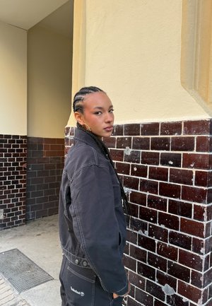 Jonge vrouw met gevlochten haar, gekleed in een zwarte jas en broek, staat zijwaarts tegen een beige muur met donkerbruine tegels.