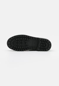 Suela de zapato de goma negra con surcos pronunciados, que presenta una puntera redondeada y una superficie texturizada para agarre y durabilidad. Logo de la marca visible.