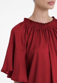 Blusa rossa con scollatura arricciata, maniche corte a campana e vestibilità morbida. Il materiale sembra morbido con una leggera lucentezza.