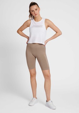 Ung kvinde står med hænderne i siden iført hvid ærmeløs cropped top med teksten "HUMMEL" og beige cykelshorts med tekst på låret.