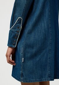 Giacca di jeans con piping bianco a contrasto sui polsini, dotata di bottoni neri. Il tessuto è di un blu medio con una texture liscia.
