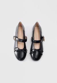 Zwarte leren Mary Jane schoenen met enkelbandjes en kleine strikken op de ronde neuzen, vanuit bovenaf getoond op een effen achtergrond.