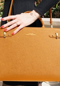 Oranje handtas met een gestructureerde afwerking, goudkleurige hardware accenten en logo. Hand met rode nagels die de tas vasthoudt. Eenvoudig, elegant ontwerp.