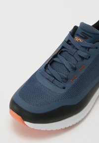 Chaussure de sport bleu foncé avec dessus en maille, accents noirs, détails orange, bout arrondi et semelle texturée pour une meilleure adhérence. Conception à lacets incluse.