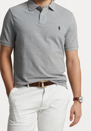 Graues Poloshirt mit Kragen und kurzen Ärmeln, versehen mit einem kleinen schwarzen Logo auf der Brust. Kombiniert mit weißen Hosen und einem braunen Gürtel.