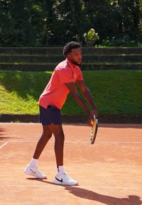 Tennisspiller i rød polo og marineblå shorts, som forbereder seg på å serve på en leirbane, holder en gul og svart racket, og har på seg hvite sko.