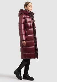 Femme portant un long manteau matelassé brillant bordeaux avec une capuche, associé à des leggings noirs et de grosses bottes noires montantes.