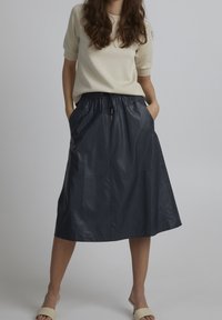 Jupe mi-longue bleu marine en matériau brillant, avec une taille élastique et des poches latérales ; portée avec un haut de couleur claire et des sandales.