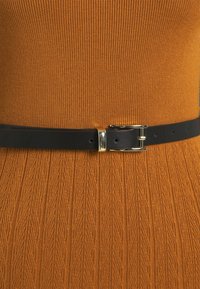 Cintura in pelle nera con una chiusura in metallo dorato, indossata sopra un top in tessuto texturizzato a coste di colore senape con un motivo a foglie.