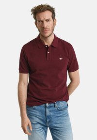 Bordeaux polo shirt met een platte kraag, korte mouwen en een borstlogo. Heeft een gladde textuur en is gecombineerd met lichtblauwe denim jeans.