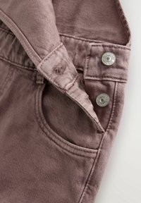 Gros plan sur un jean en denim rose montrant une poche avant et une ceinture avec deux boutons métalliques étiquetés "Next Jeans Since 1982."