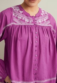 Lilla bomuldsbluse med broderede hvide blomsterprint, rund halsudskæring, samlet skæring og knaplukninger foran.
