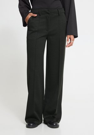Femme portant un pantalon noir à jambes larges et des chaussures noires, vêtue d'un haut noir à manches longues avec une main dans la poche.