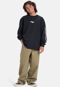 Svart langermet skjorte med et grafisk logo på brystet og hvit tekst på ermene, kombinert med løse beige bukser og svarte joggesko.