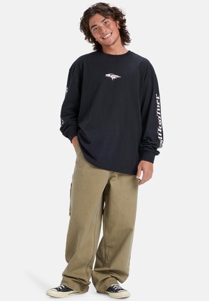 Schwarzes Langarmshirt mit einem grafischen Logo auf der Brust und weißem Text an den Ärmeln, kombiniert mit lockeren beigen Hosen und schwarzen Turnschuhen.