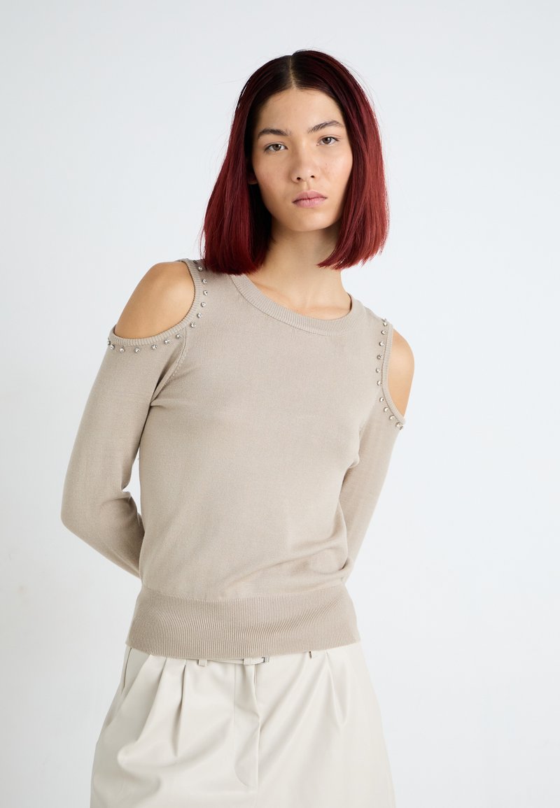 Maglione beige a maglia con design a spalla fredda, impreziosito da borchie metalliche lungo le cuciture delle spalle e un orlo a costine aderente.