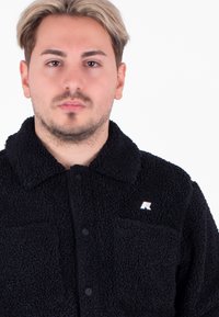 Zwarte fleece jas met een kraag, twee voorzakken en een klein logo op de linkerborst. Zachte textuur en knoopsluiting.