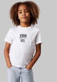 Weißes Baumwoll-T-Shirt mit kurzen Ärmeln, mit einem schwarzen Grafikdruck, der "CALVIN KLEIN 1978" zeigt. Kombiniert mit hellblauen Jeans.