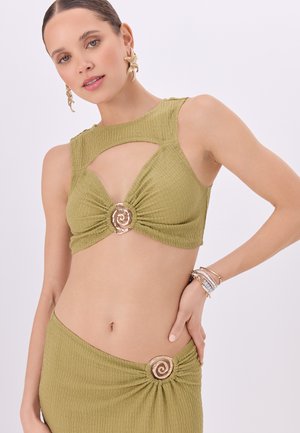 Vrouw die een olijfgroene getextureerde cropped top en bijpassende rok draagt met spiraalvormige gouden accenten, gecombineerd met armbanden en grote bloemenoorbellen.