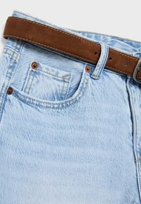 Jeans in denim azzurro chiaro con tasca frontale visibile, rivetti marroni e cintura in suede marrone infilata nelle passanti.