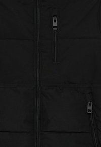 Svart polstret jakke med glidelås foran og to gliselommer. Jevn tekstur med horisontale sømmer og slitesterkt stoff.