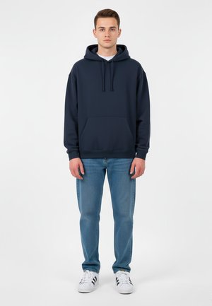 Felpa navy con tasca frontale, cappuccio con cordino e polsini a coste, abbinata a jeans light blue e sneakers bianche con dettagli neri.