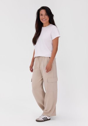 Frau mit langen dunklen Haaren, lächelnd, trägt ein weißes T-Shirt, beige Cargohosen und weiße Sneaker mit schwarzen Streifen, stehend auf weißem Hintergrund.