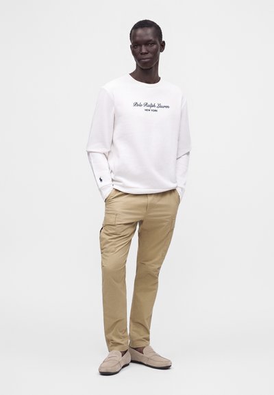 Άνδρας που φοράει λευκό πουκάμισο Polo Ralph Lauren με μακριά μανίκια, μπεζ παντελόνια cargo και ανοιχτό καφέ loafers, στέκεται με τα χέρια στις τσέπες.