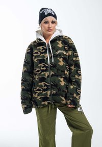 Giacca in pile camouflage verde e beige con chiusura con zip, colletto alto e dettagli ricamati, abbinata a pantaloni verde oliva. Berretto nero in testa.
