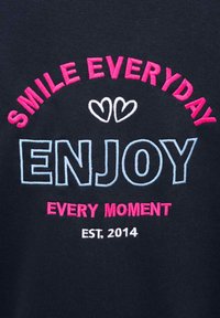 Granatowa tkanina z haftowanym napisem: „UŚMIECHAJ SIĘ CODZIENNIE” w różowym, „CIESZ SI�Ę” w jasnoniebieskim oraz „KAŻDĄ CHWILĄ” w różowym, z dwoma sercami powyżej.