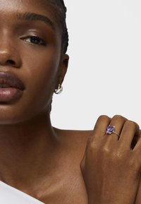 Anillo de plata con una gran gema púrpura, llevado en una mano con piel marrón texturizada. Diseño minimalista, en un fondo blanco.