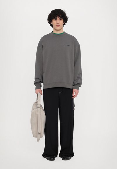 Grå oversized sweatshirt med ribbede mansjetter, sammen med svarte vidde bukser. Lys grå skulderveske holdt med én hånd. Glatt stofftekstur.