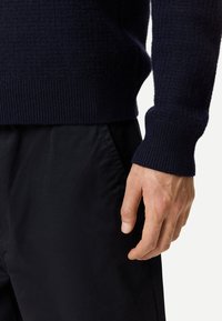 Marinblå stickad tröja med ribbade muddar och fåll; bärs över svarta byxor. Tröjan har ett strukturerat mönster och mjukt tyg.