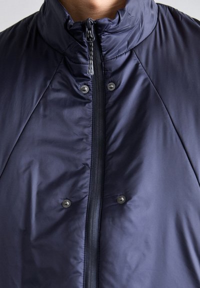 Giacca imbottita blu navy con colletto alto, chiusura frontale con zip e bottoni a pressione. Presenta una texture liscia e lucida e un motivo trapuntato.