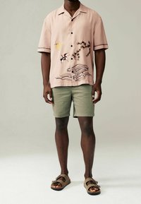 Jasnoróżowa koszula z krótkim rękawem z czarnymi guzikami i misternym haftem z wizerunkiem żurawia, słońca oraz fal. Zestawiona z oliwkowymi szortami i brązowymi sandałami.