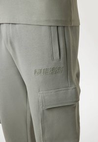 Jasnozielone cargo dresy o teksturowanym wykończeniu; cechy to szwy kieszeni oraz wytłoczone logo "GUESS" na boku.