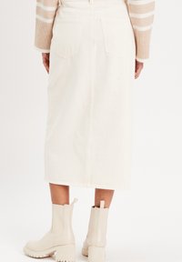 Falda midi de mezclilla blanca con bolsillos traseros, que presenta una silueta recta. Combinada con botines beige.