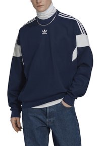 Mörkblå sweatshirt med vita detaljer, rund halsringning och oversized passform. Har Adidas-logotypen på bröstet och tre ränder på axlarna.