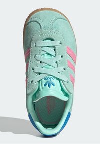 Světle zelená semišová teniska s růžovými pruhy, strukturovanými šňůrkami a gumovou podrážkou. Na patě je modrý akcent a logo Adidas.
