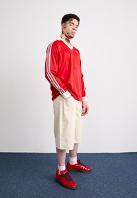 Rød atletisk langermet skjorte med hvite striper, beige shorts og røde joggesko med svarte detaljer, på en person som står på et blått teppe.