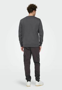 Homme aux cheveux bouclés vu de dos, portant un sweat-shirt gris foncé, un pantalon cargo noir et des baskets blanches, sur un fond uni.